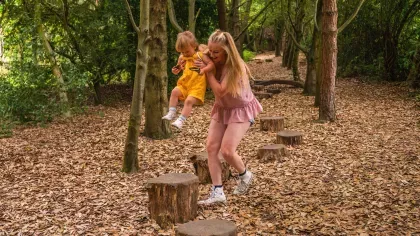 A blonde mother swings a toddler child along a line of tree stumps small enough to use as stepping stones