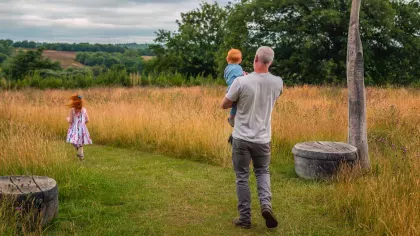 A man is carrying a child into a meadow, a girl is running ahead.