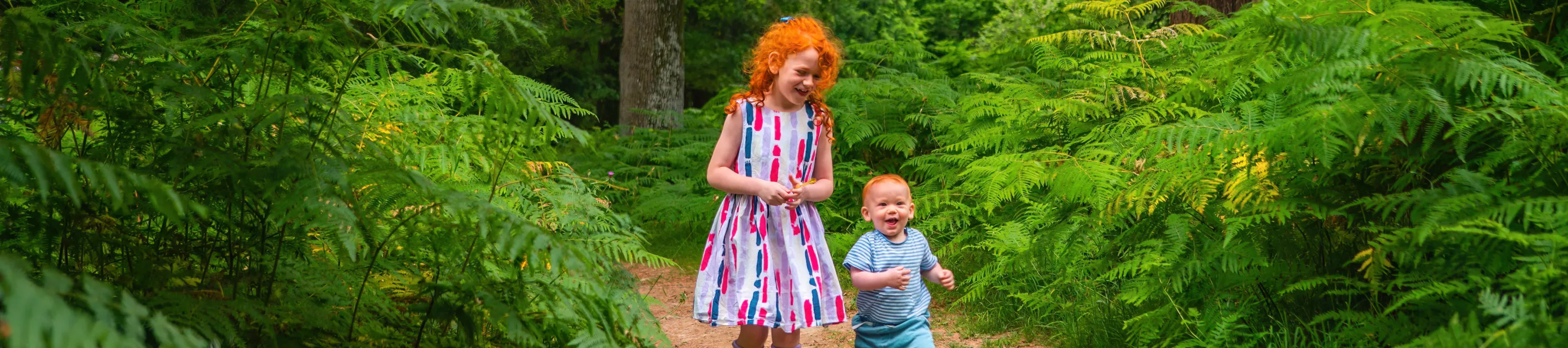 A girl with red curly hair and a toddler boy walking through green shady woodlands