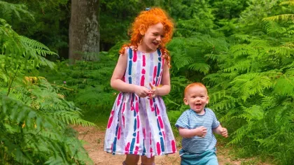 A girl with red curly hair and a toddler boy walking through green shady woodlands