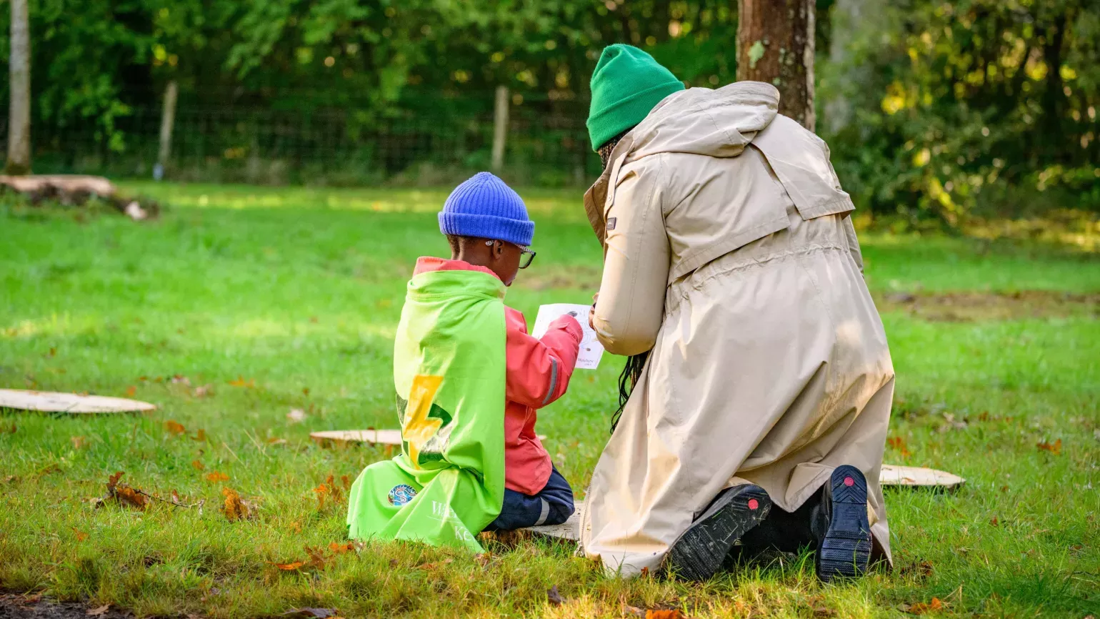 Seen from behind, a mother kneels next to her child on a grassy lawn at the start of a trail of stepping stones, both of them looking at a sheet of paper