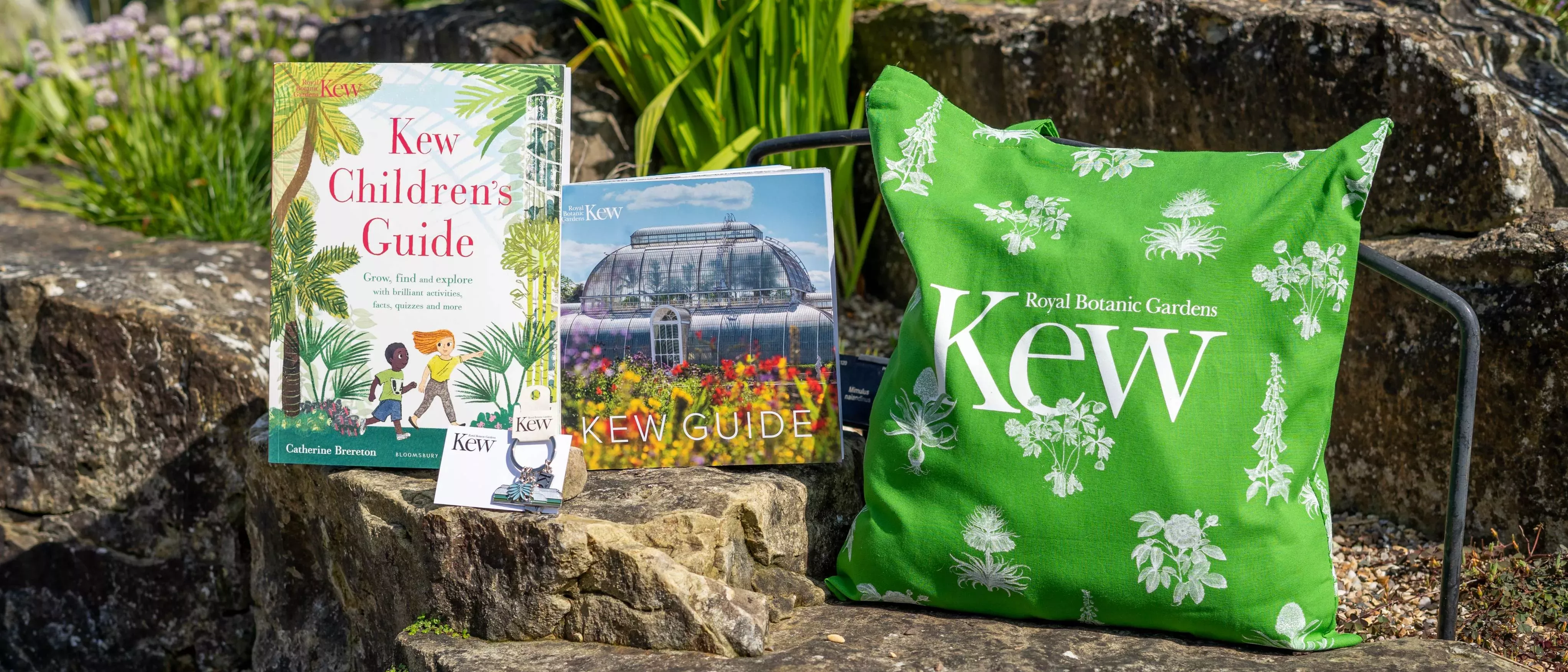 Four products displayed on a rock wall growing plants: a book titled "Kew Children's Guide", a book titled "Kew Guide", a keyring featuring glasshouse, palm leaf and Kew logo pendants, and a green tote bag with a Kew logo and plant prints in white