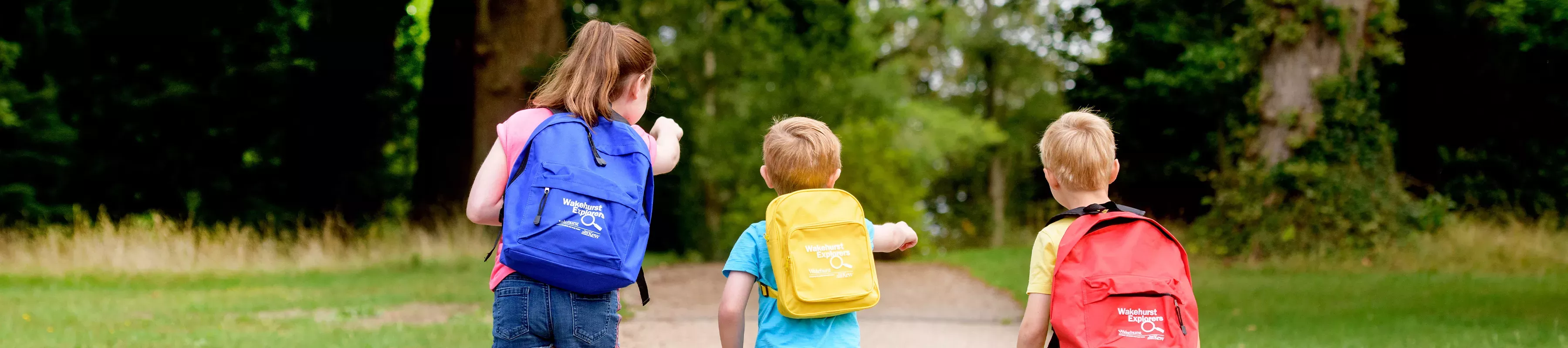 Three children walk down a forest path, each with a Wakehurst Explorer rucksack