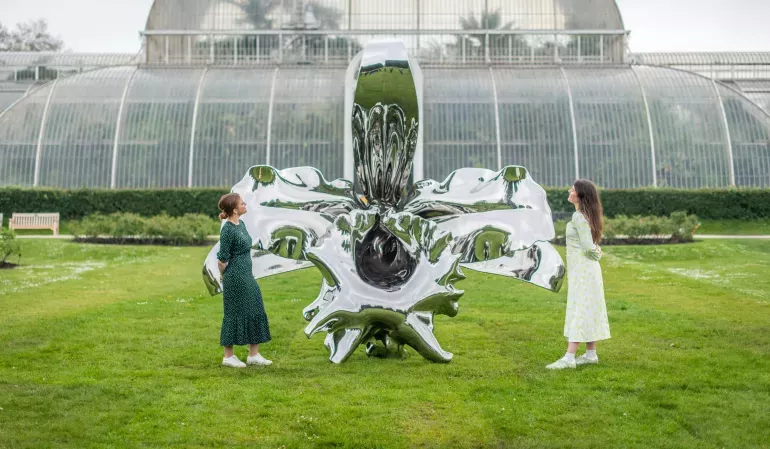 Two young women stand on either side of a giant shiny metal sculpture of an orchid flower, on a lawn in front of a Victorian glasshouse