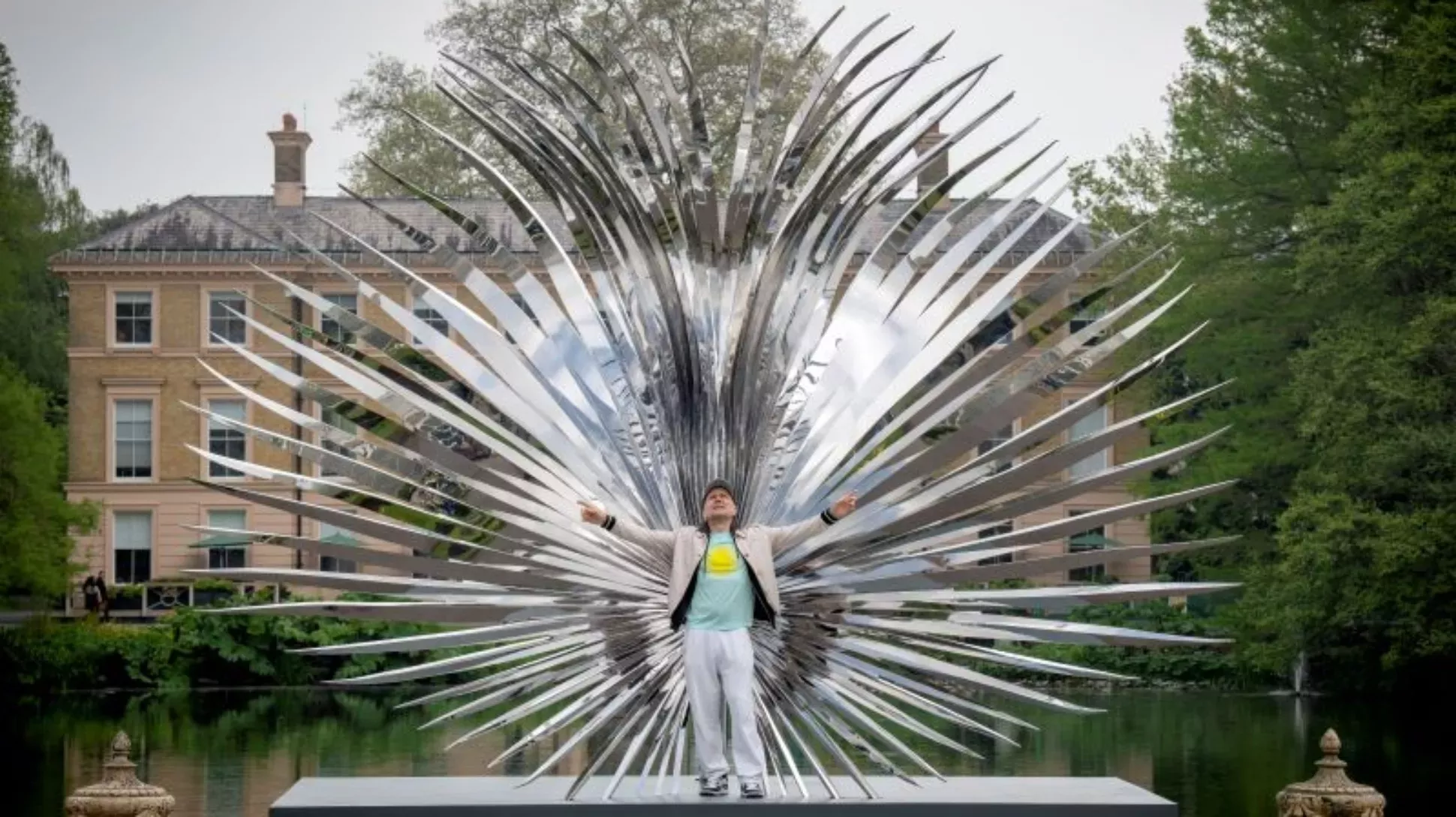 Marc Quinn standing in front of a large shiny metal palm leaf sculpture at Kew Gardens