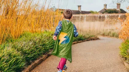 A child runs through a garden at Wakehurst wearing a superhero cap with a lightning bolt on it