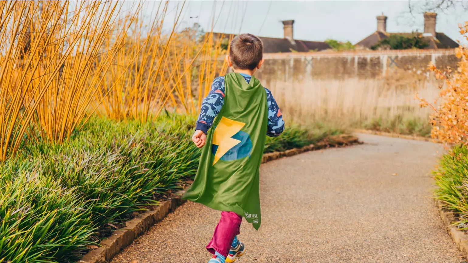 A child runs through the Winter Garden at Wakehurst with a green Nature Heroes cape