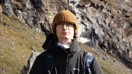 A person wearing a yellow hat and black coat in front of a rocky arctic landscape