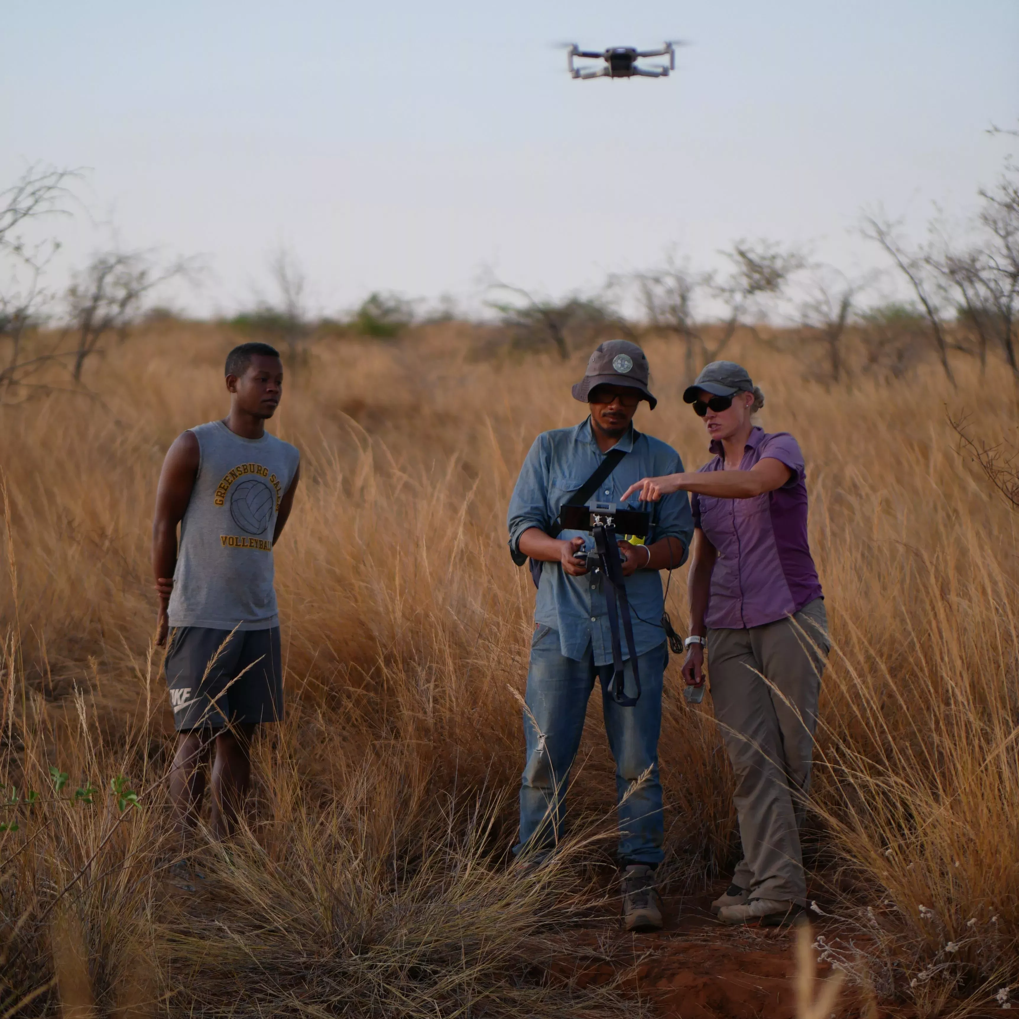 People stand in an area of grassland, one of them pilots a drone which hovers nearby