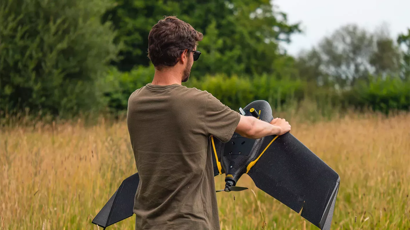 A scientist stands in a field holding a fixed wing drone