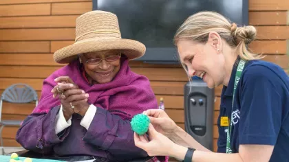 Elderly Black woman in a straw hat smiling at a young white woman in volunteer uniform, who is examining a tropical fruit moulded in teal modelling clay