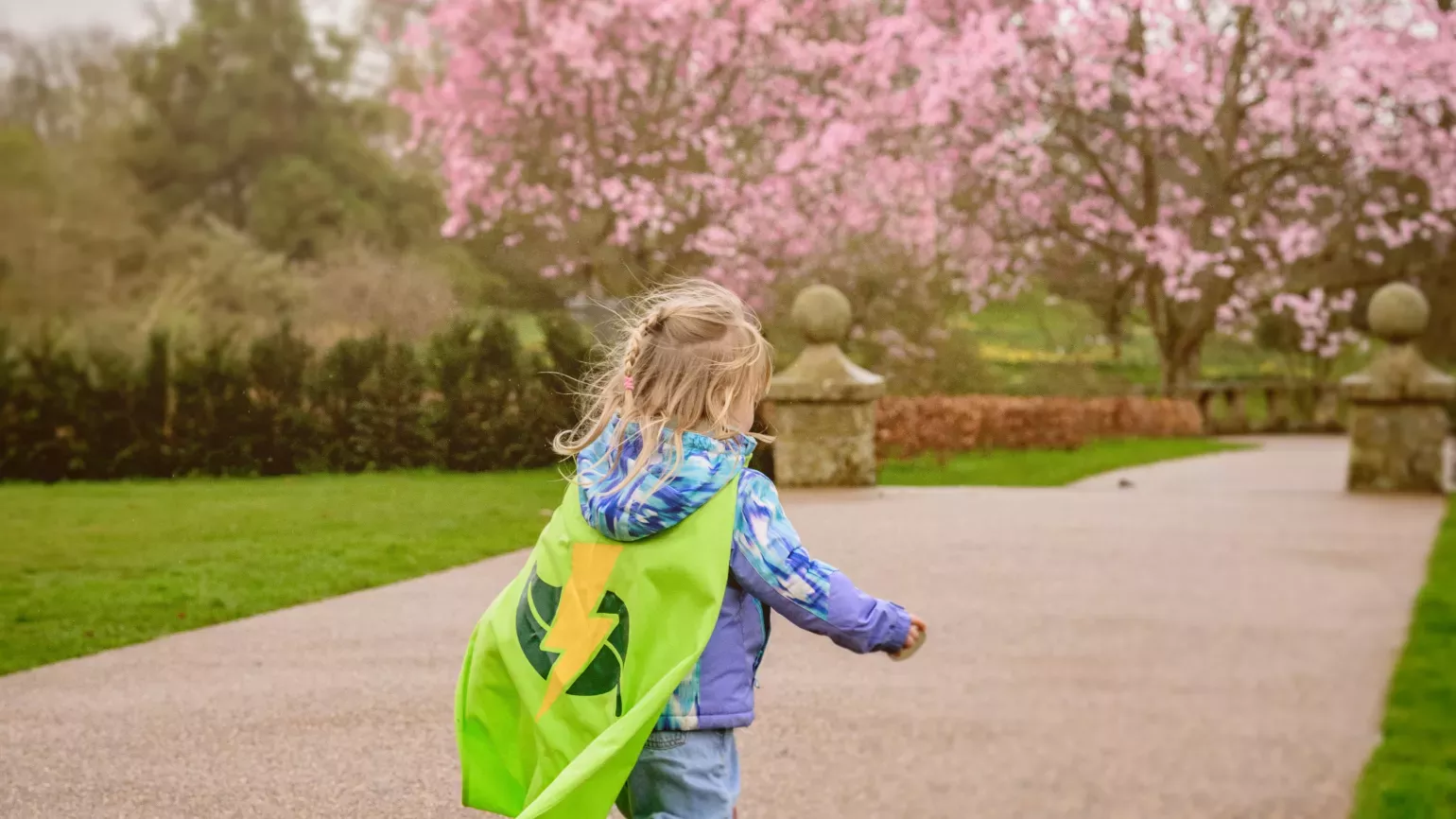 A small girl in a parka, wellies and green Nature Heroes cape runs down a garden path towards cherry trees in bloom