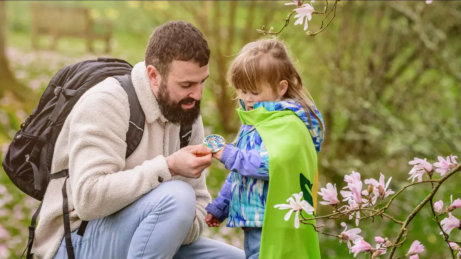 A young girl in a green Nature Heroes cape examines a badge in her hand with her dad, a bearded man in outdoor clothing kneeling next to her. A green park is behind them and pink magnolia blossoms are in the foreground