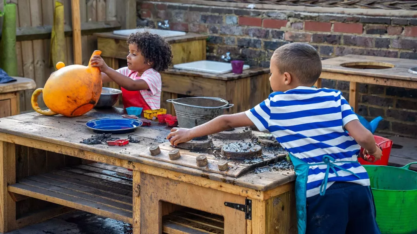 A young boy leans over a wooden oven, whilst a girl holds up a plastic watering can.