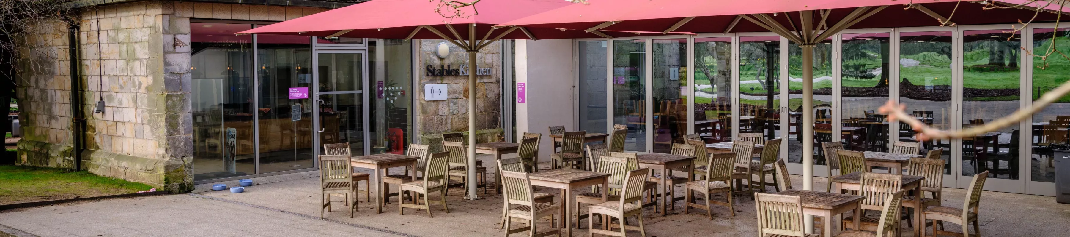 Wooden tables and chairs under red umbrellas outside Wakehurst's Stables Kitchen