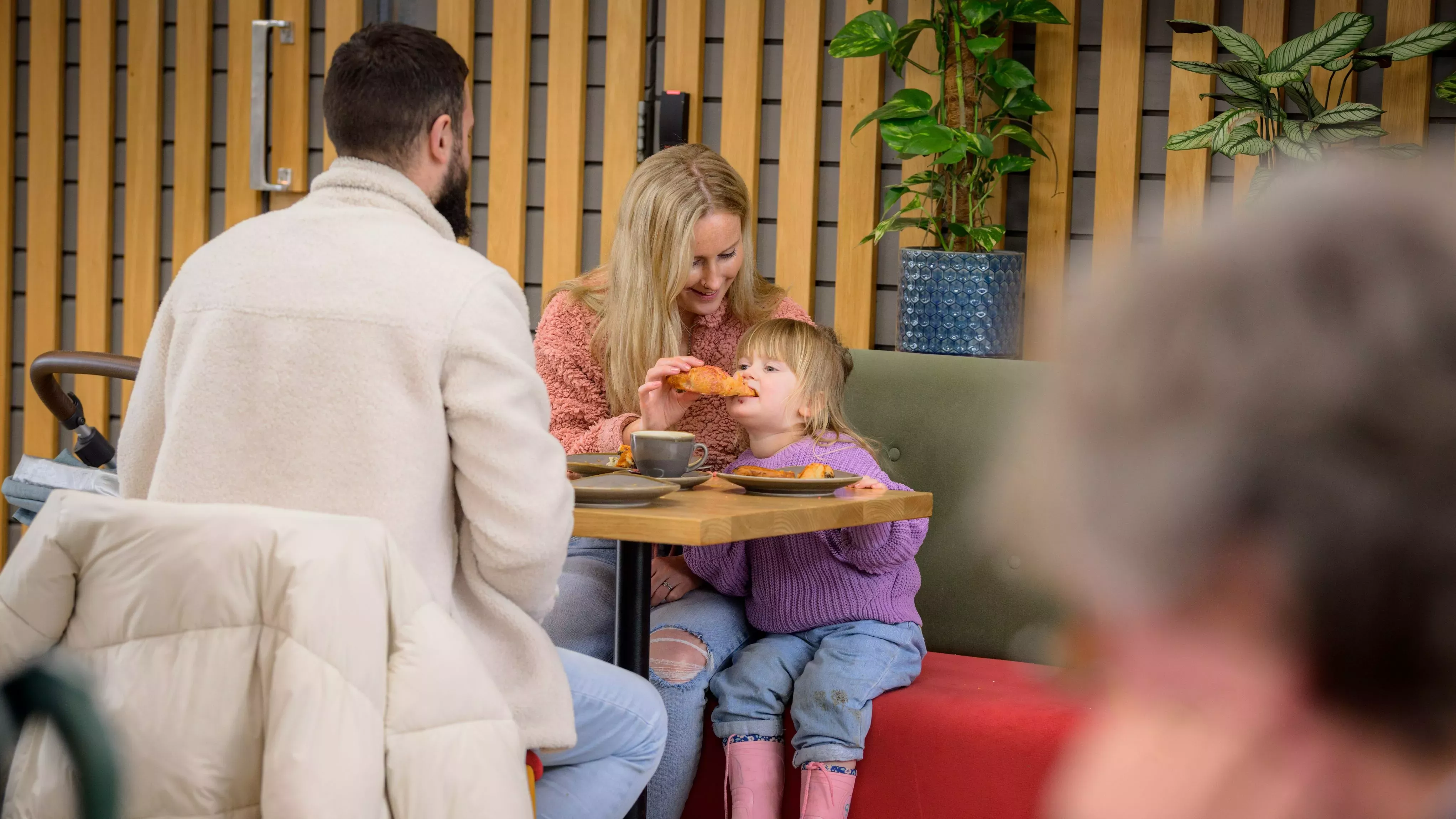 A mother feeds a pastry to her young daughter in a cosy cafe while dad looks on